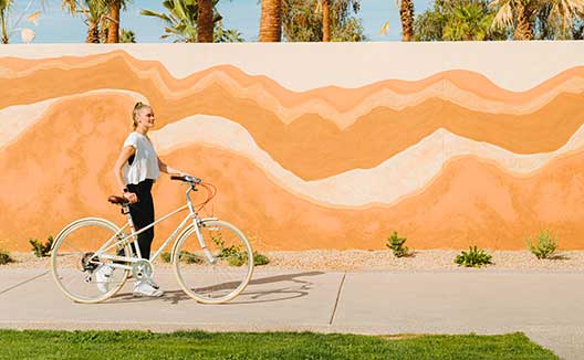 woman walks with bicycle in front of a painted wall at Kimpton miralina resort